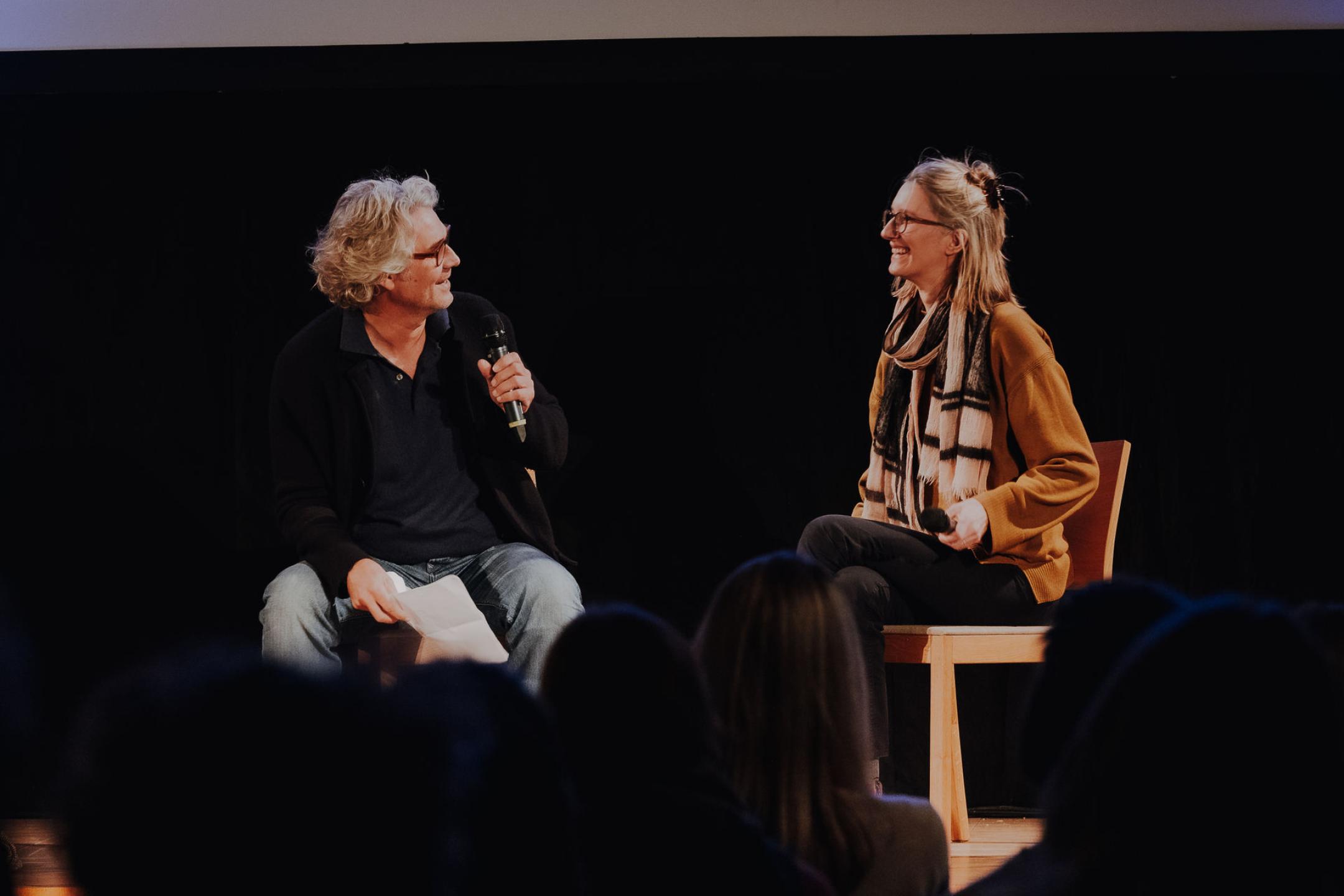 Sitting on chairs on a stage, to people are talking and laughing with each other