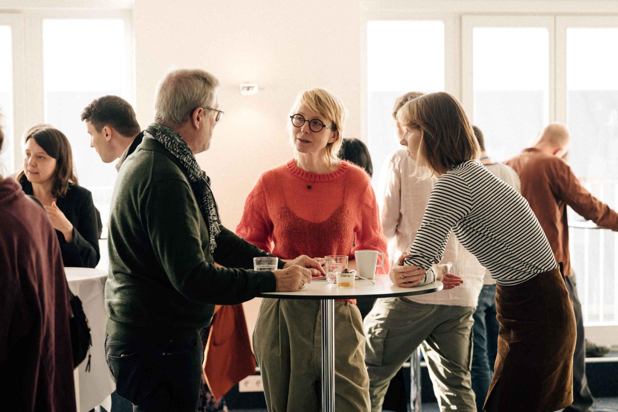 Three people in conversation at a standing table, behind them are more people busily talking to each other