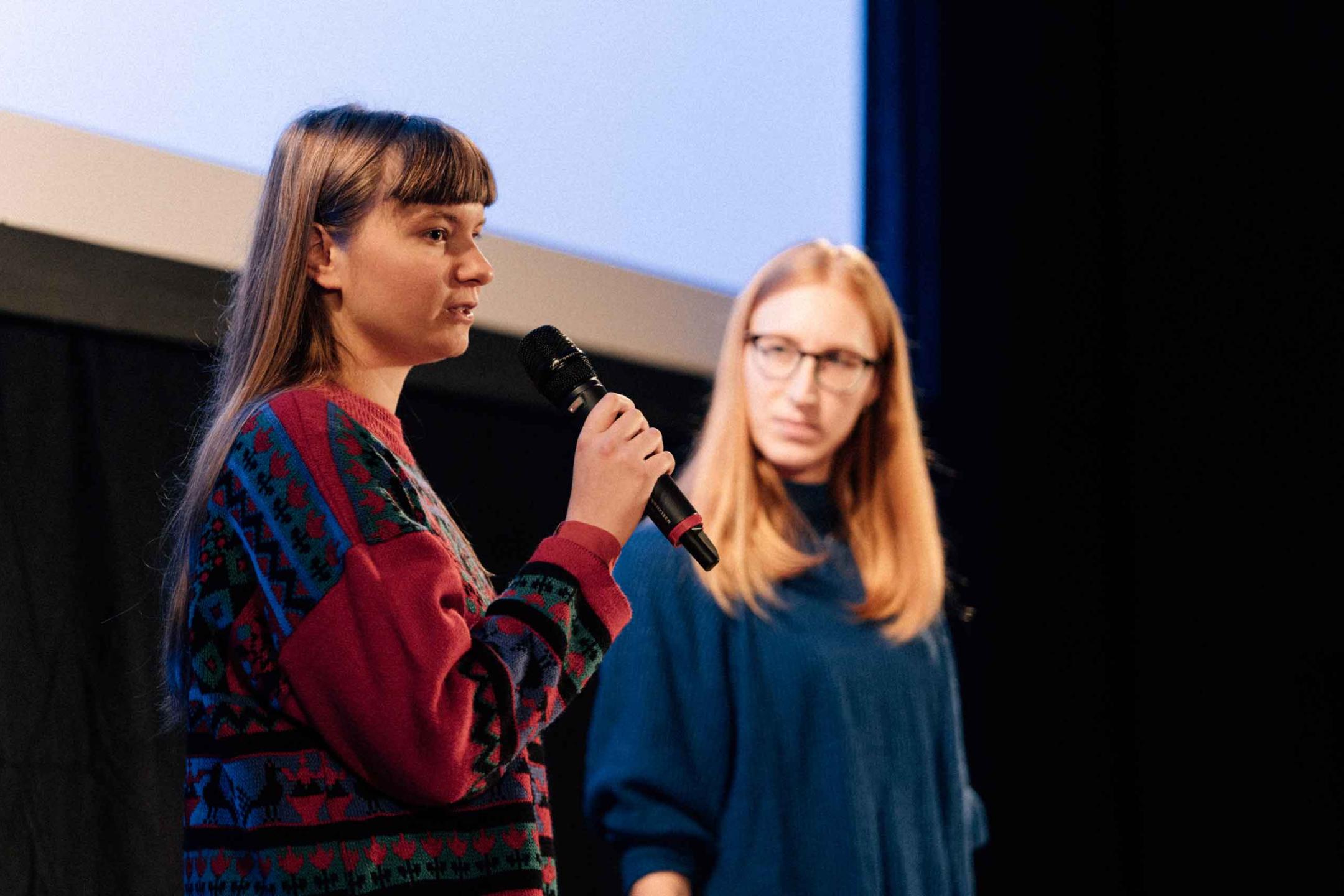 A woman on stage is speaking into a microphone, the woman next to her listens intently