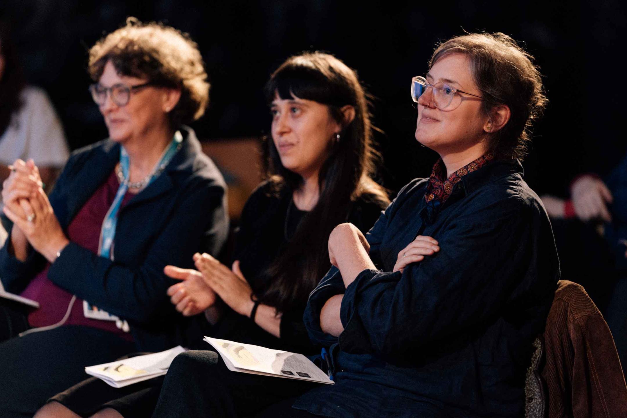 Three panelists are looking kindly towards a stage an begin to clap their hands