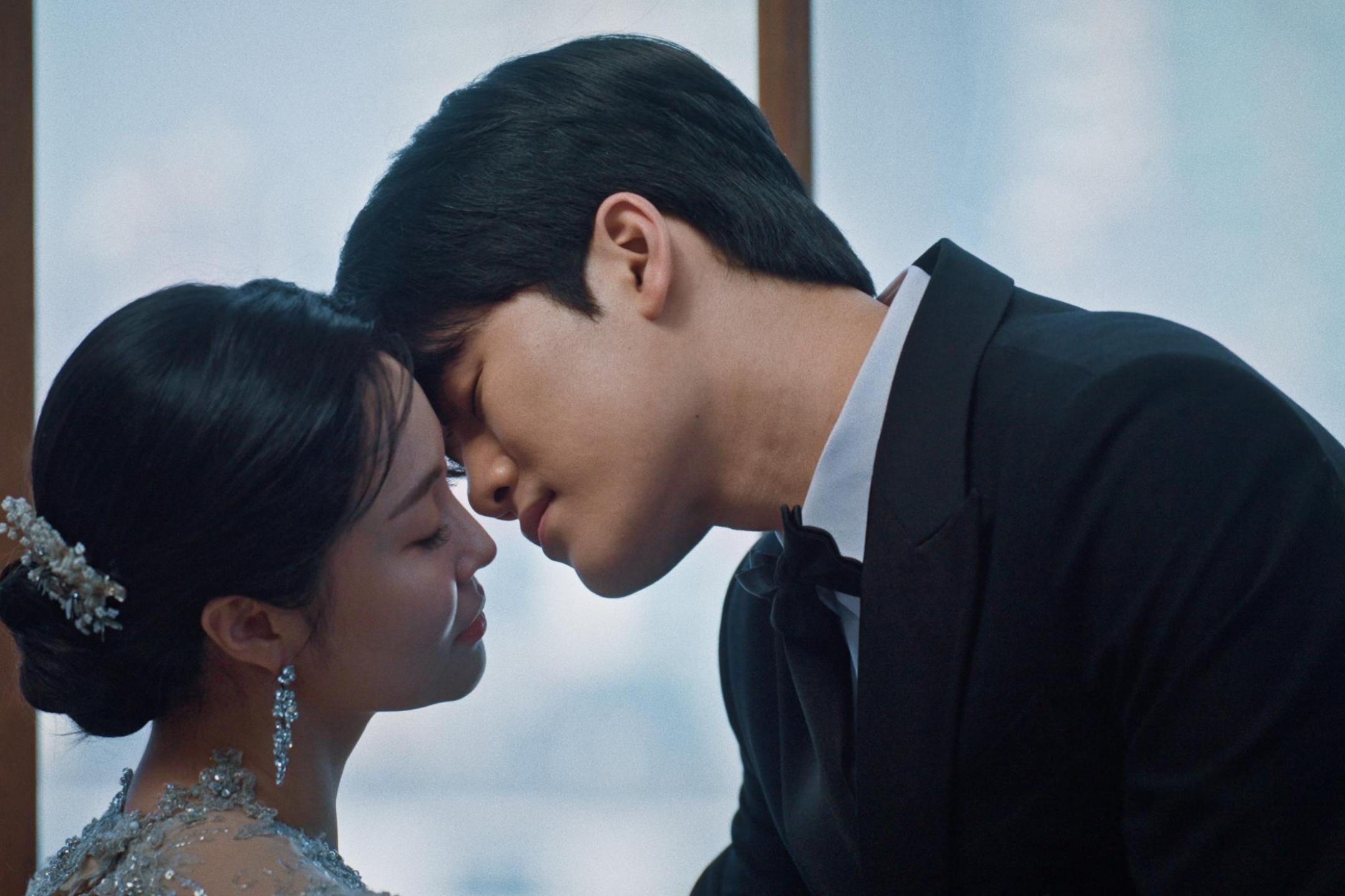 Close up on a Korean man in a suit leaning towards a Korean woman in a sparkly dress to kiss her