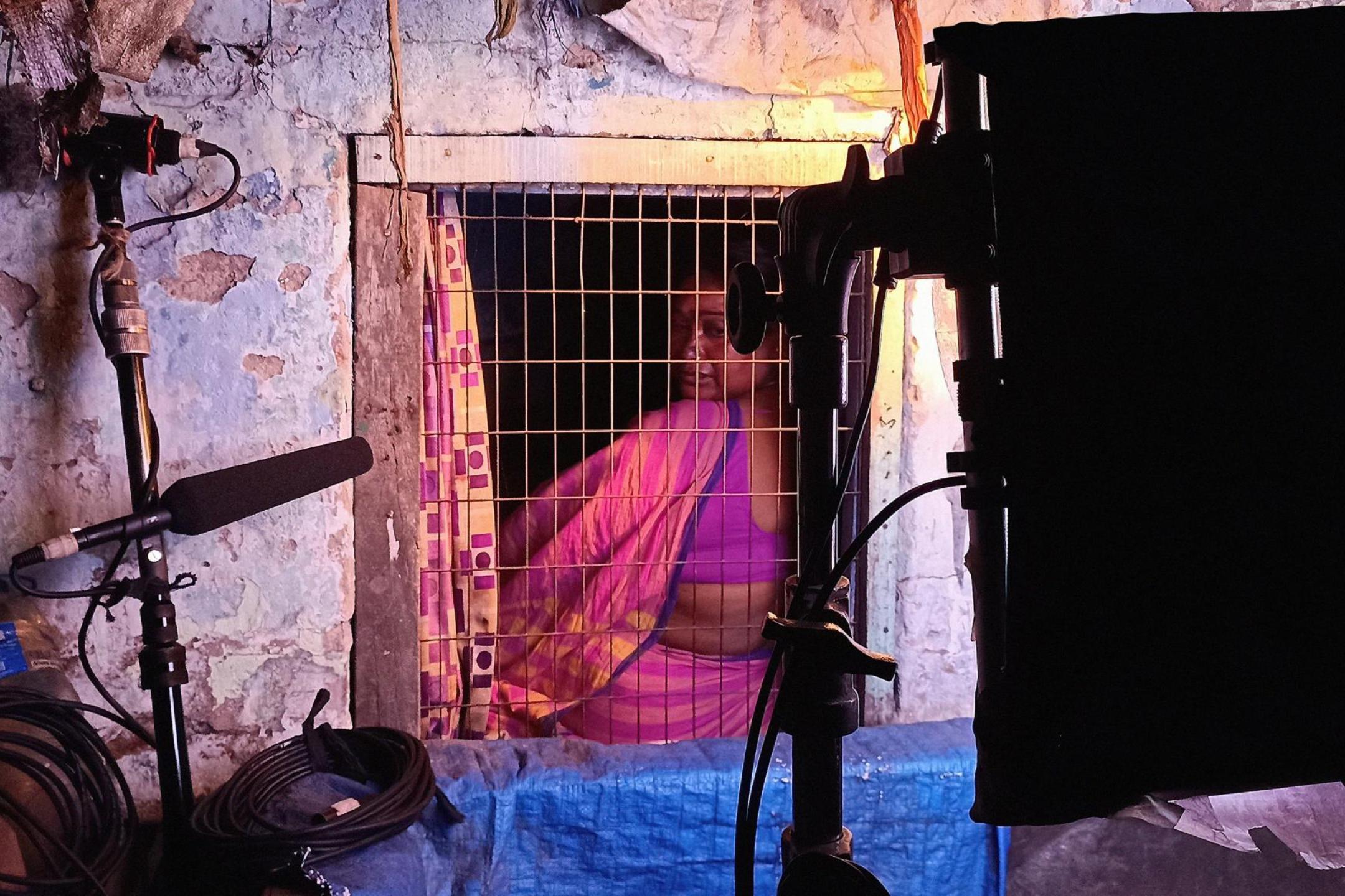 A woman wearing a sari is sitting behind a barred window and looks over her shoulder to the camera and the filmset in front of the window