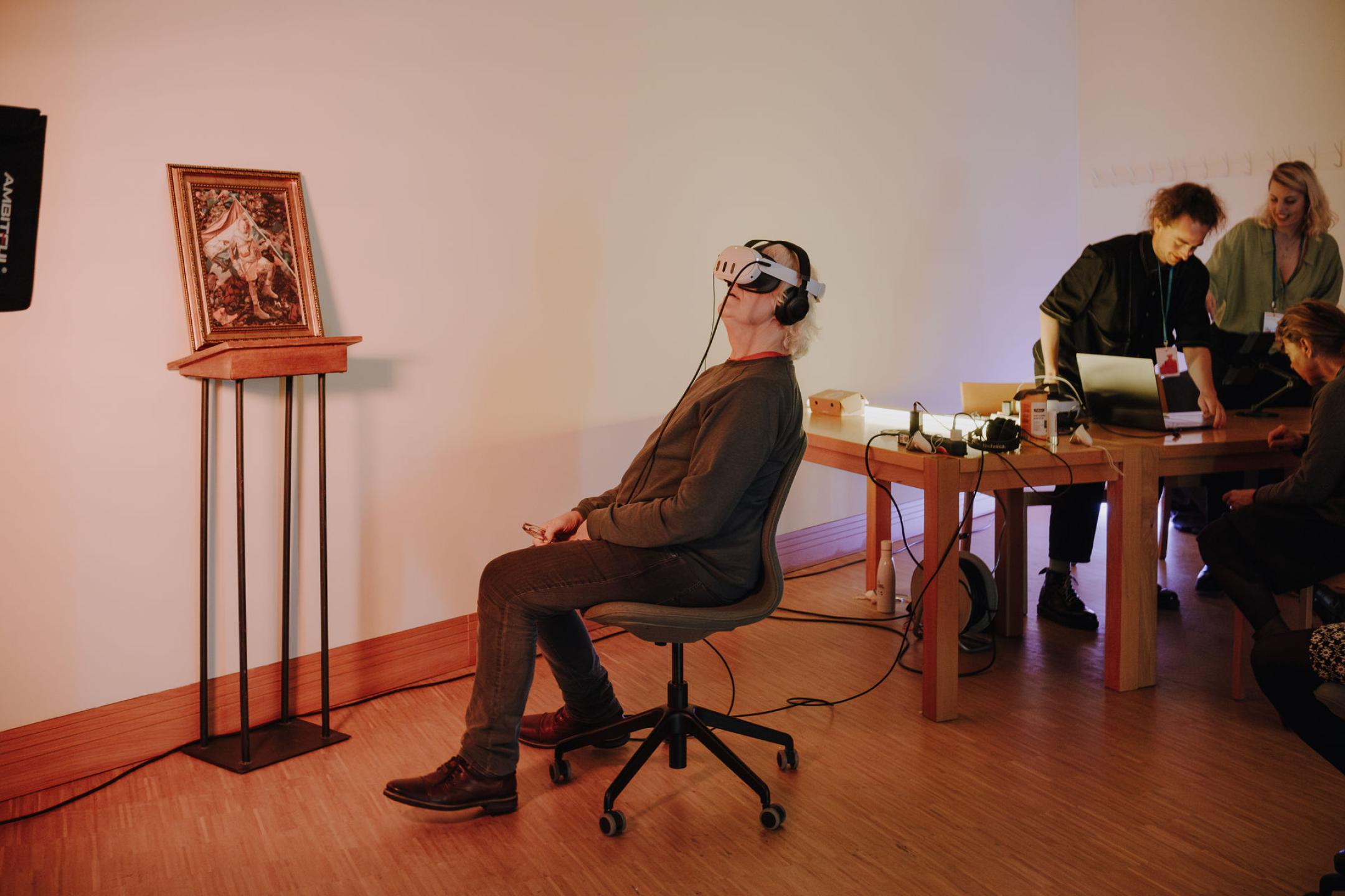 An older person is sitting on a swiveling chair wearing vr glasses and headphones. next to him is a painting in a gold frame and on his other side, people are gathering around a laptop