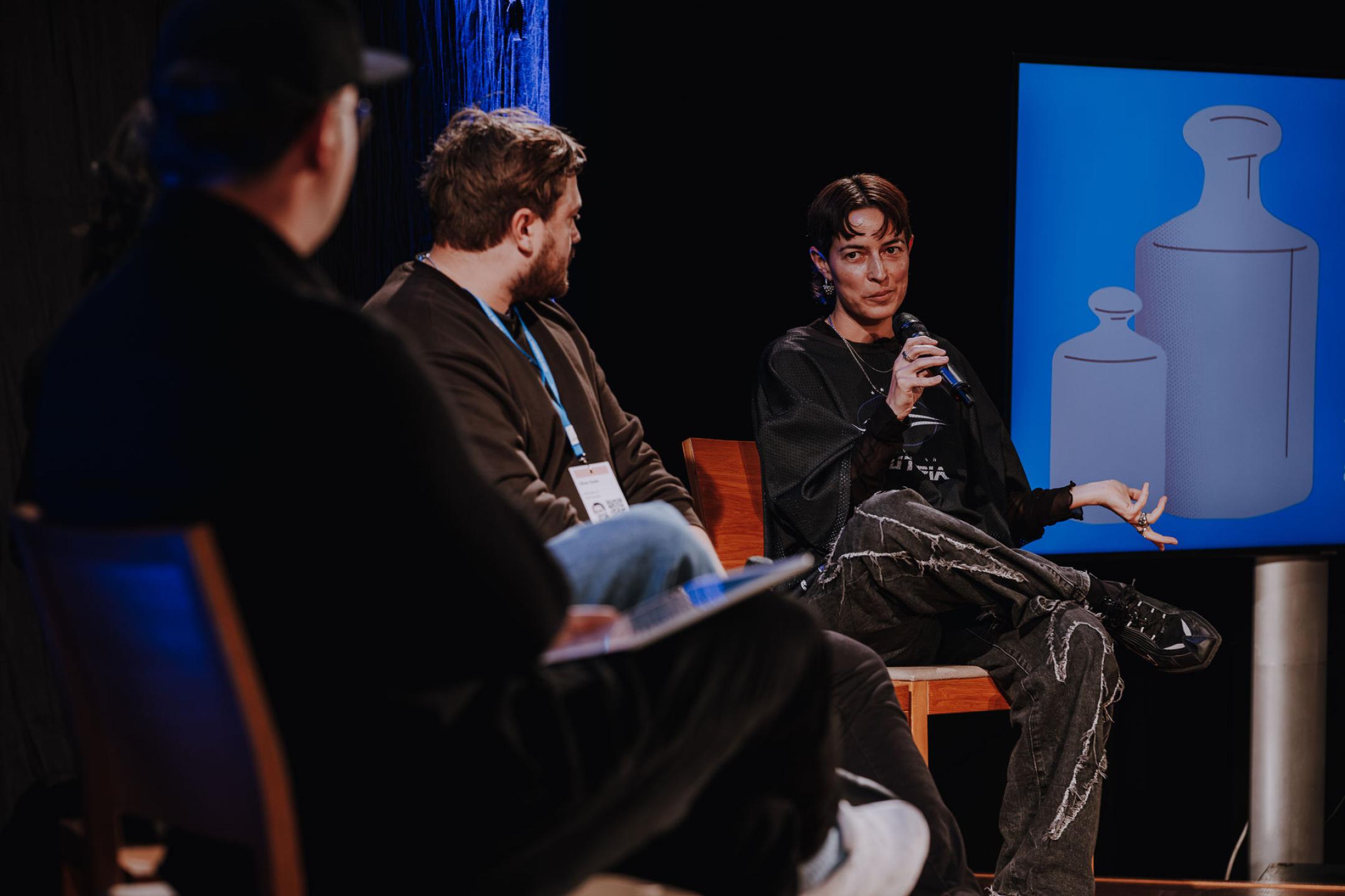 A panel with three people on stage, one person talks into a microphone and gesticulates