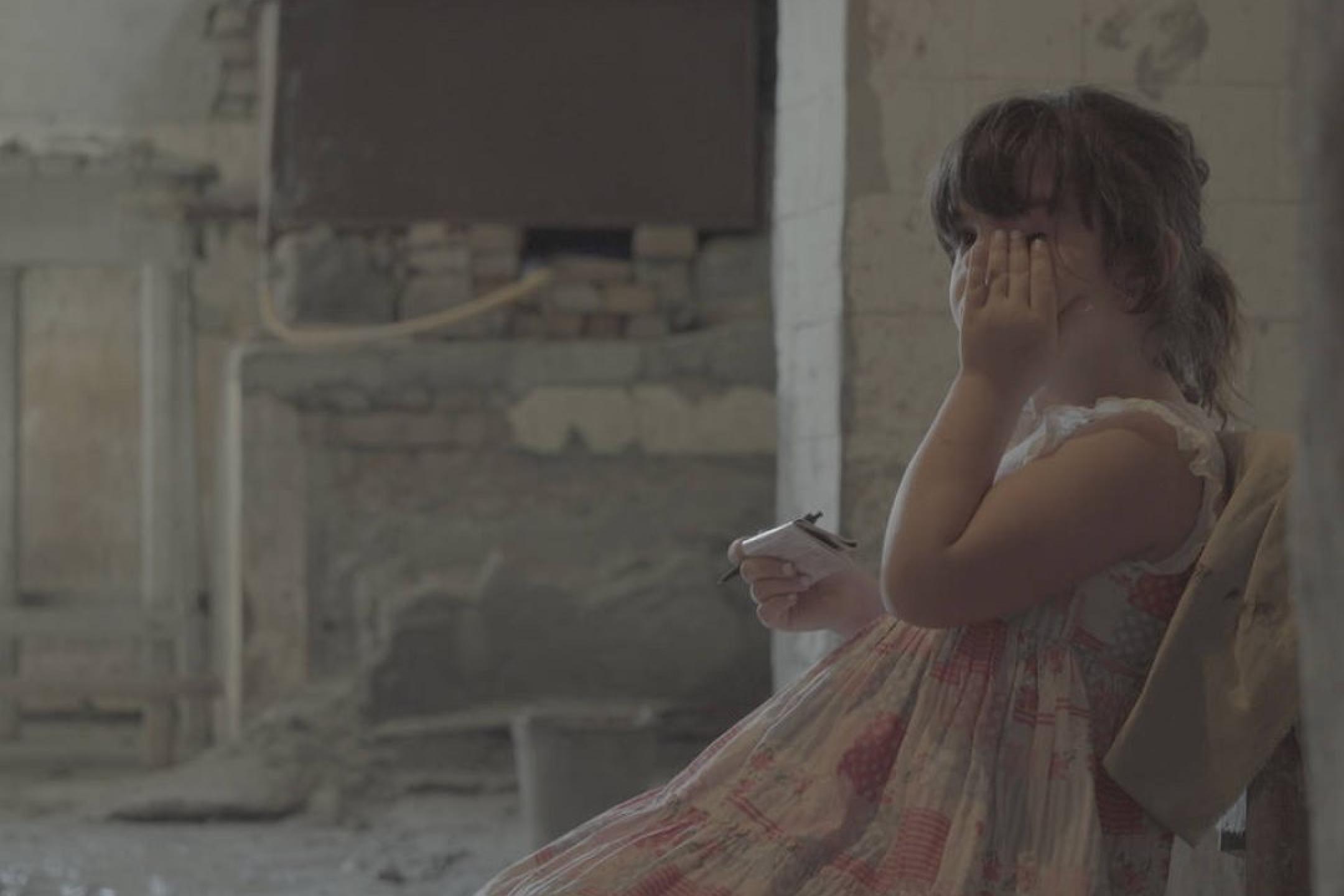 A young child in a dress is sitting on a chair in the midst of a construction side, it is rubbing her eye and holding a small paper pad and a pen