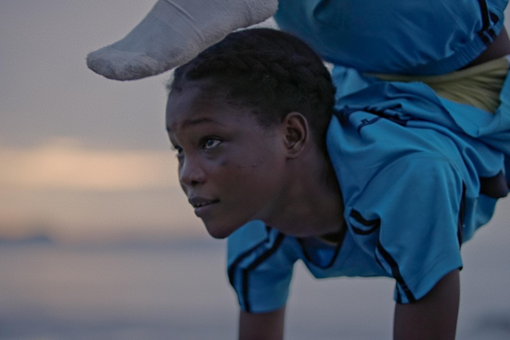 Ein junges Schwarzes Mädchen balanciert auf ihren Händen, ihr Körper ist dabei so verdreht, dass ihr Fuß beinahe ihren Kopf berührt, sie schaut konzentriert