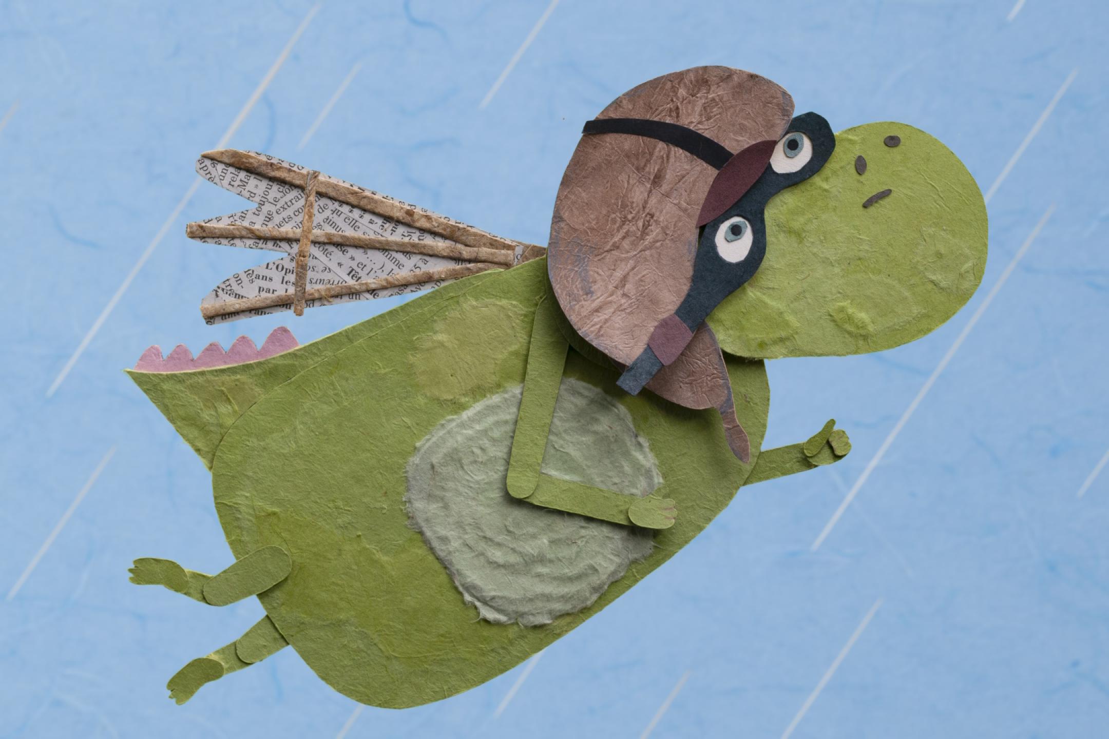 Ein grüner, gebastelter Drache mit Fliegermütze und -brille fliegt durch den Pappmaschee-Regen