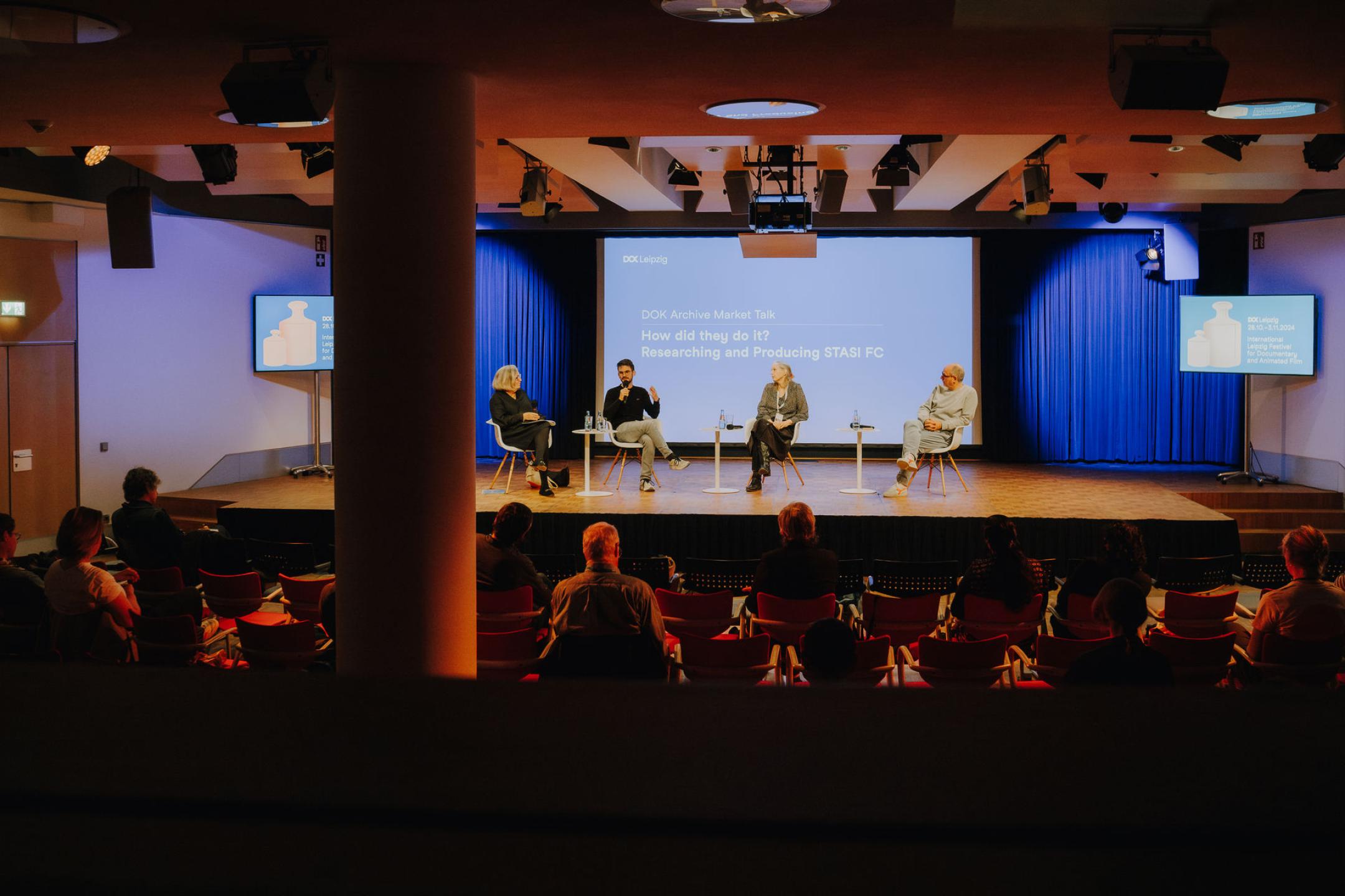 Vier Personen sitzen auf Stühlen auf einer Bühne, Publikum vor ihnen
