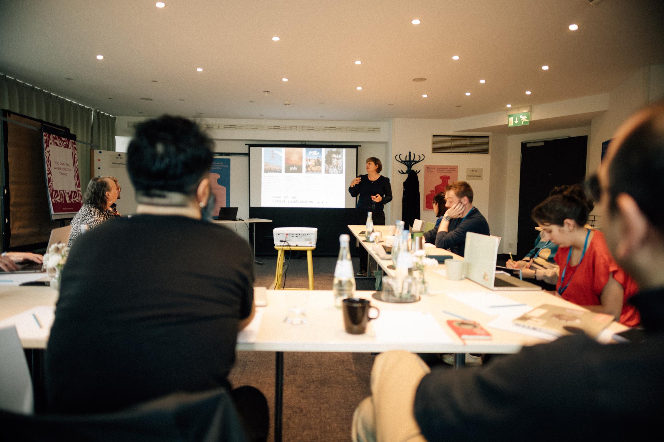 Personen sitzen an einem Tisch in einem Konferenzraum und schauen auf einen Bildschirm