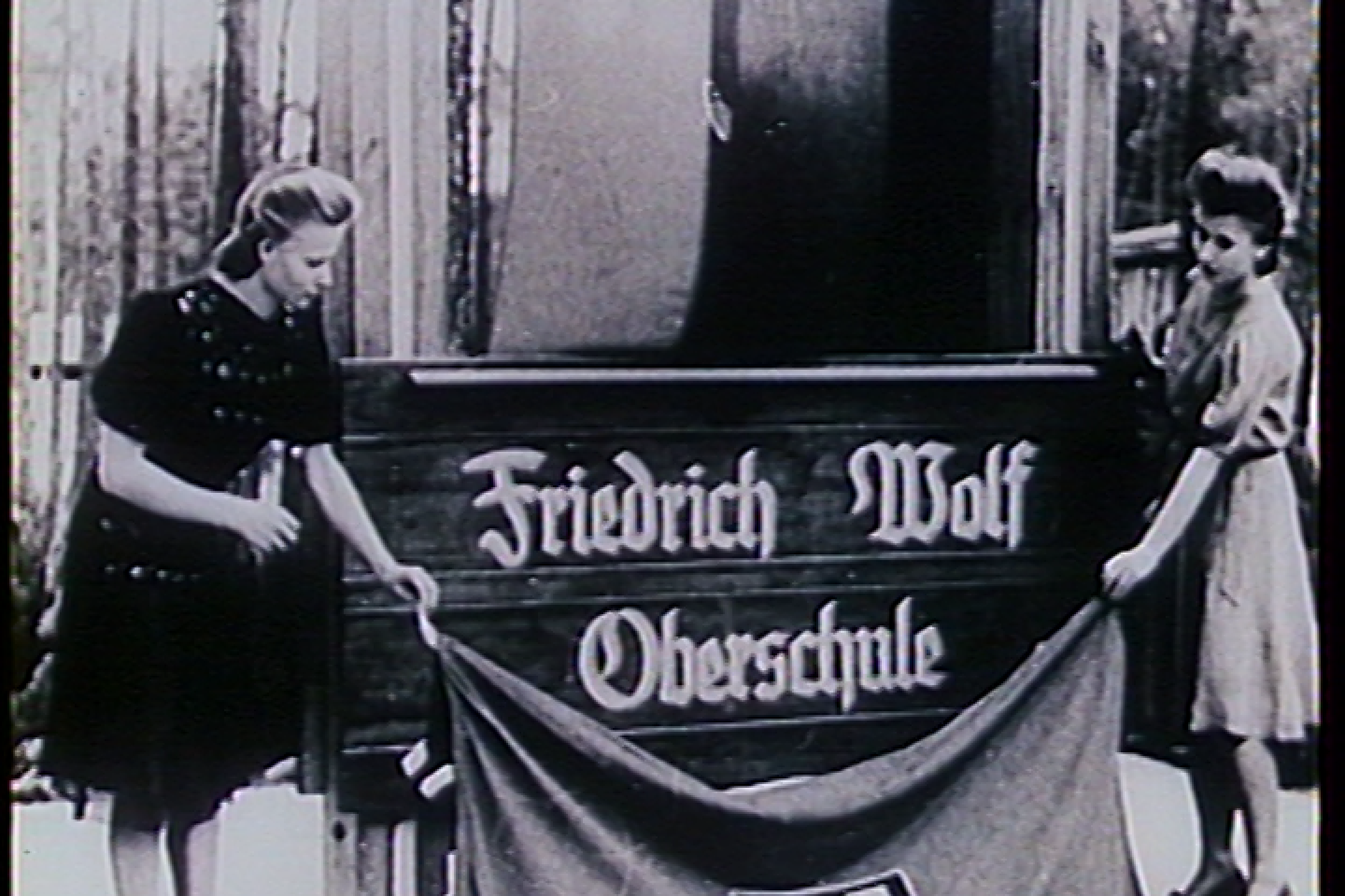 Ein analoges Filmbild, auf dem zwei Frauen in knielangen Kleidern ein Schild mit der Aufschrift "Friedrich Wolf Oberschule" enthüllen. Auf dem Stoff, den sie auf den Boden legen steht "FDJ"