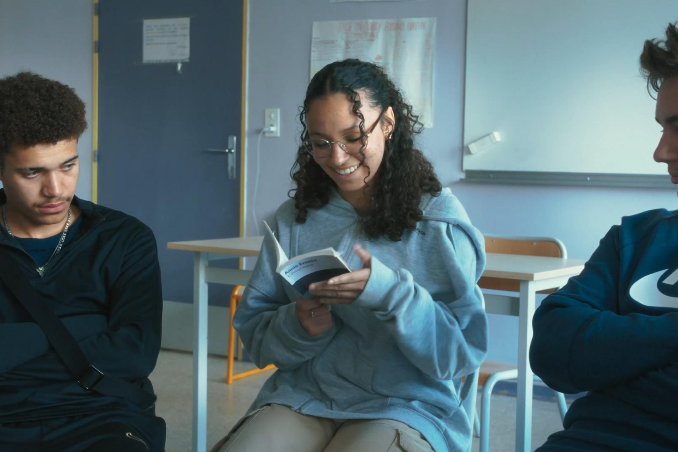 Eine Gruppe junger Menschen sitzt in einem Klassenraum im Stuhlkreis; eine Person liest aus einem Buch vor. Die anderen hören aufmerksam zu.