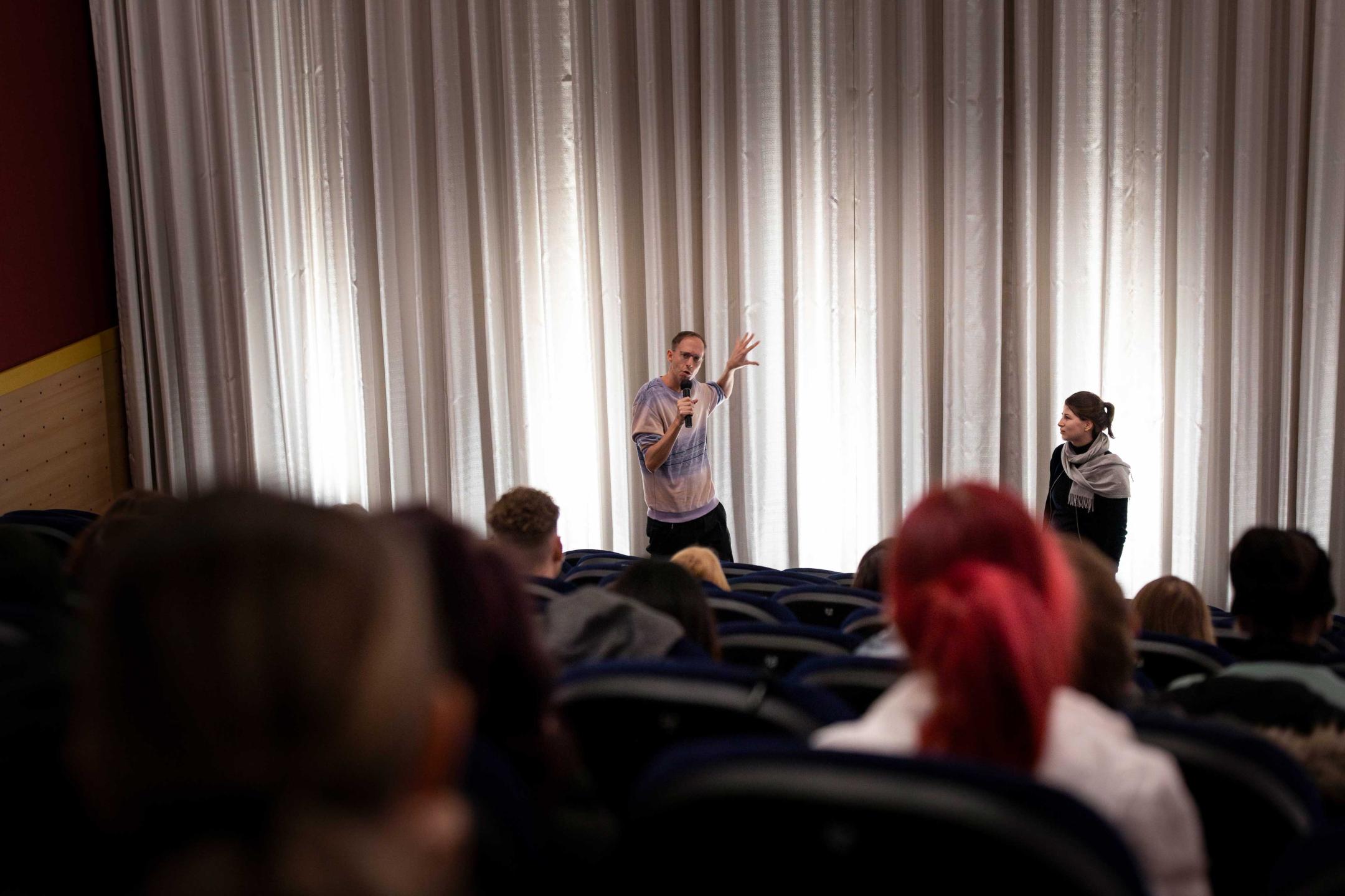 Filmregisseur Daniel Abma steht vor einem Kinopublikum und gestikuliert auf einen weißen Kinovorhang, während er in ein Mikrofon spricht