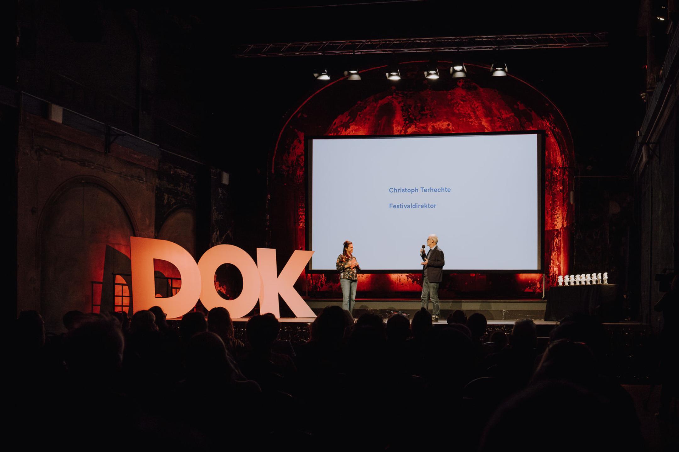 In der Schaubühne Lindenfels ist eine große Leinwand und die menschengroßen Buchstaben DOK angestrahlt. Auf der Bühne stehen vor einem vollen Publikum Christoph Terhechte, Festivalleiter und die Moderatorin Julia Weigl