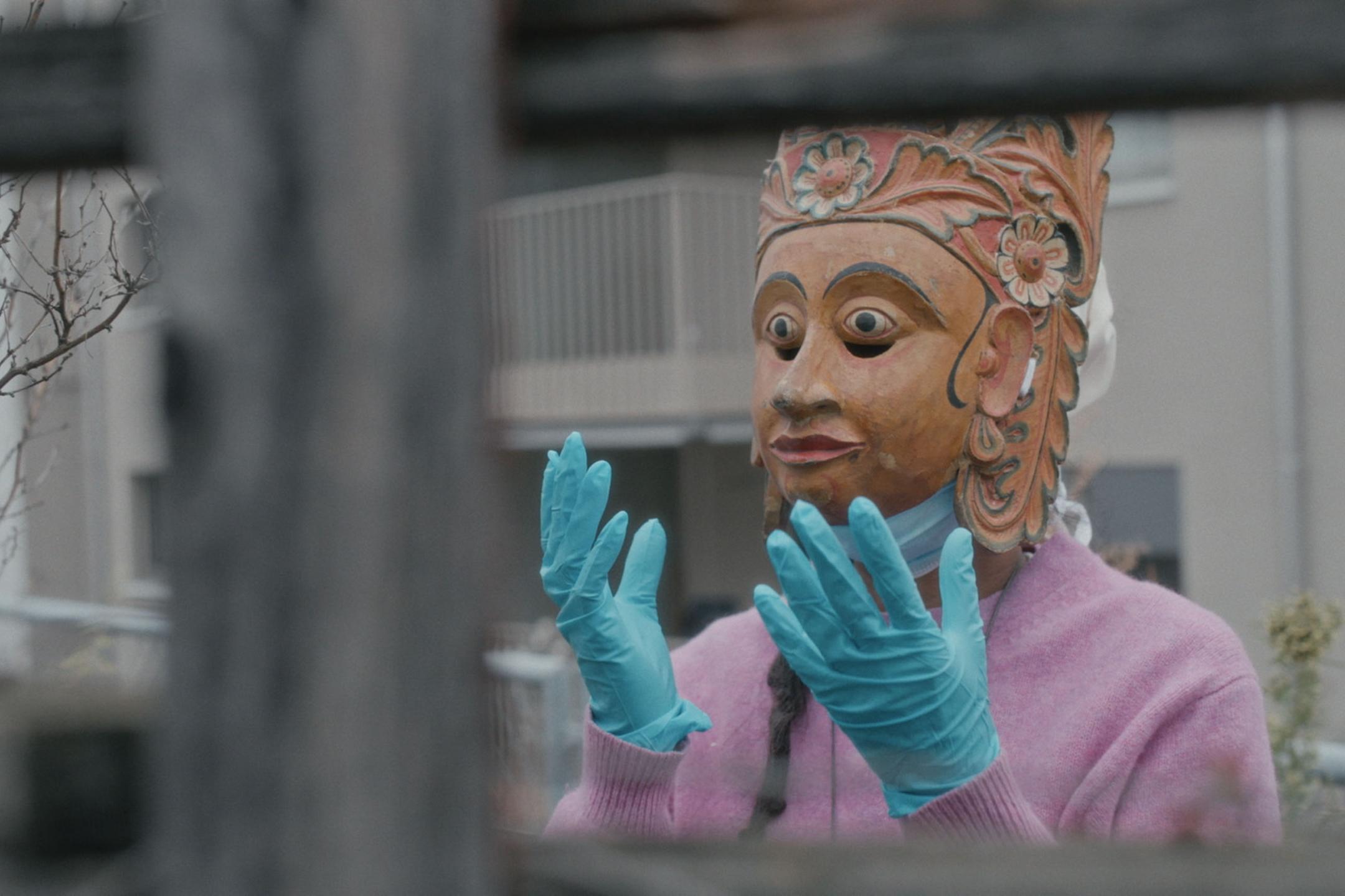 Person mit großer Holzmaske und blauen medizinischen Handschuhen