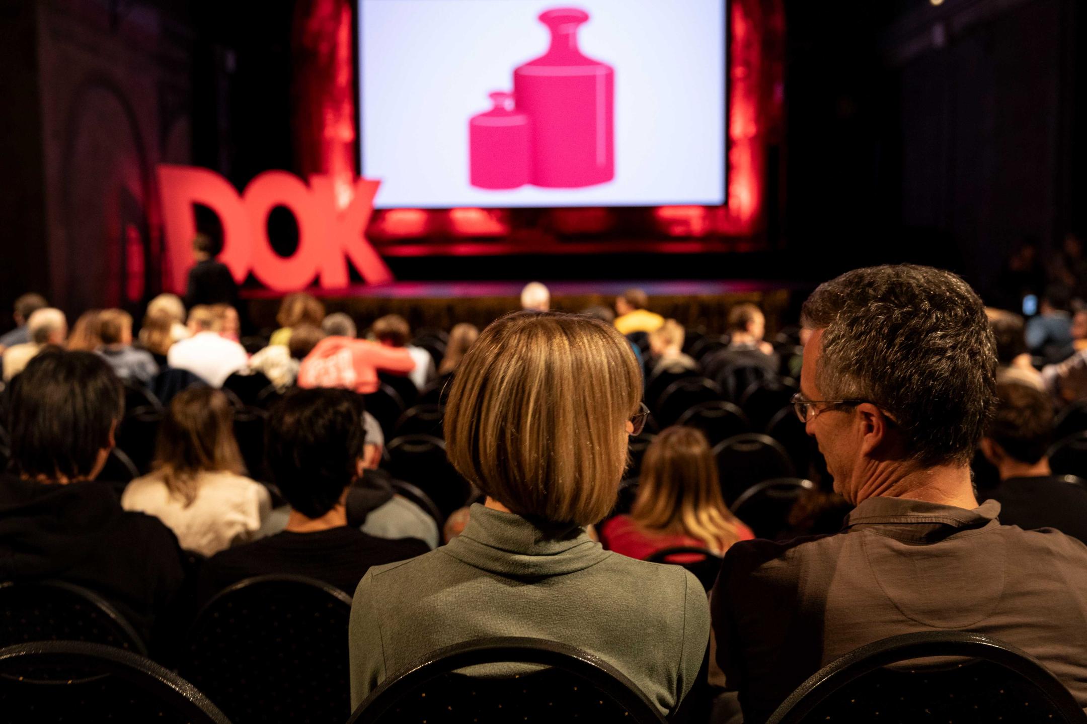 Ein Publikum in Kino, im Vordergrund schauen zwei Besucher*innen zueinander, auf der Bühne leuchtet das DOK Motiv und menschengroße DOK-Buchstaben sind auf der Bühne aufgebaut