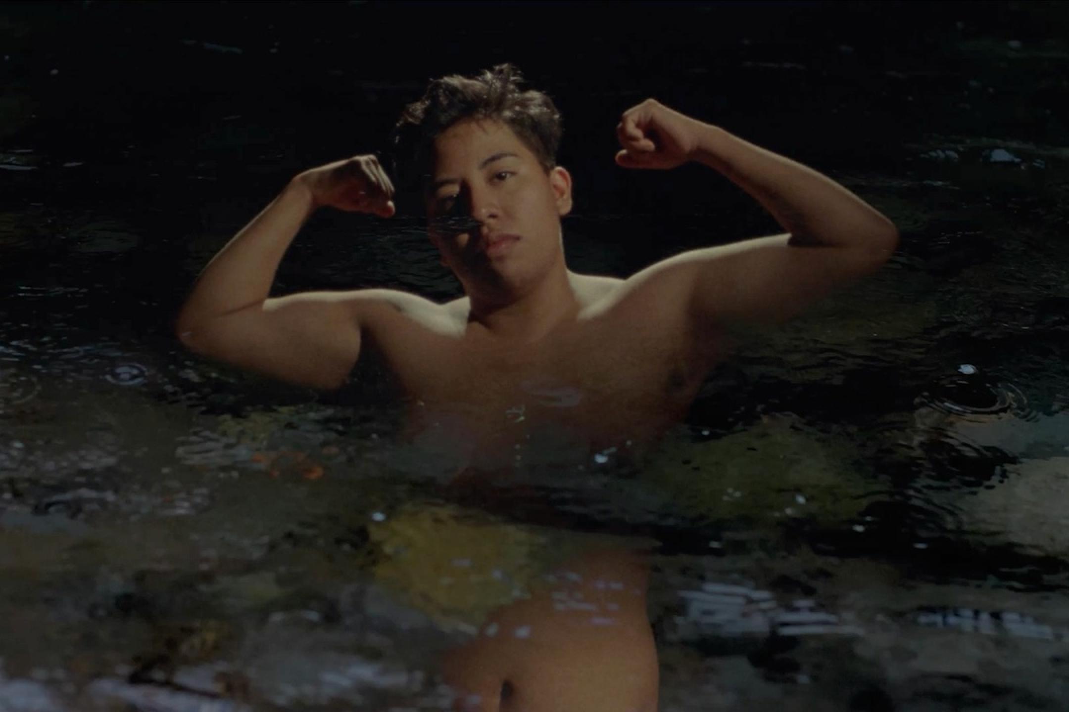 Ein Junger Mann steht brusttief im Wasser und hebt die Arme, um seinen Bizeps zu zeigen. Dabei schaut er ruhig in die Kamera