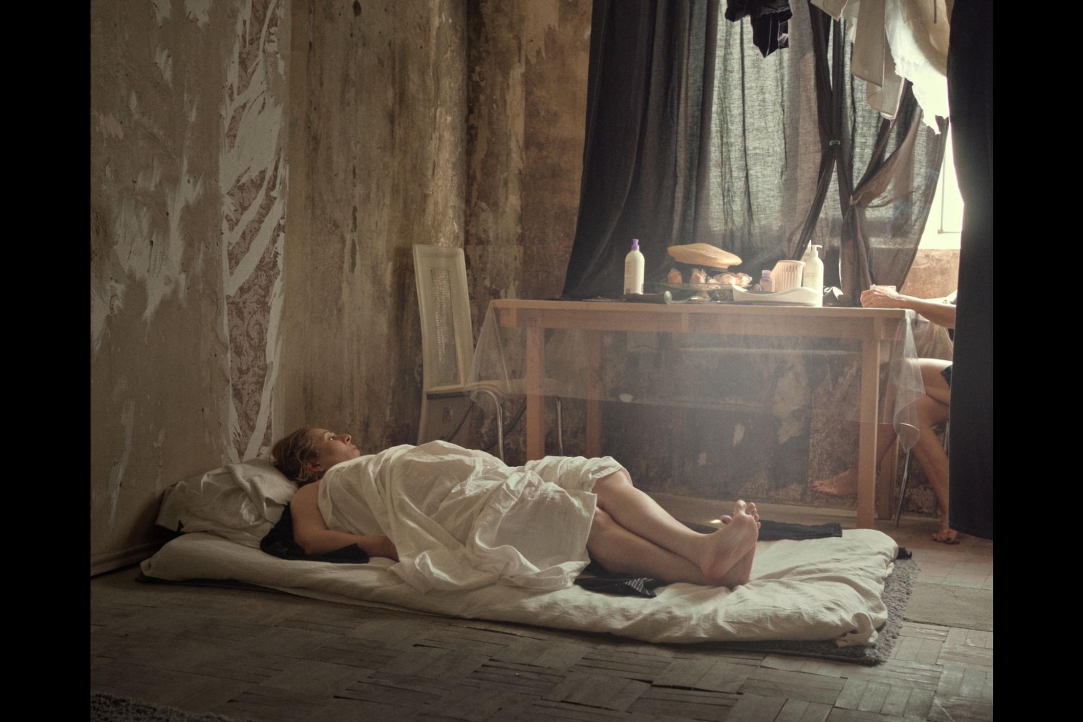 eine Frau liegt auf einer Matratze auf dem Boden, zugedeckt von einer dünnen weißen Decke. Neben ihr ein ein Tisch mit Essen und Flaschen, das Fenster ist mit Stoff verhängt