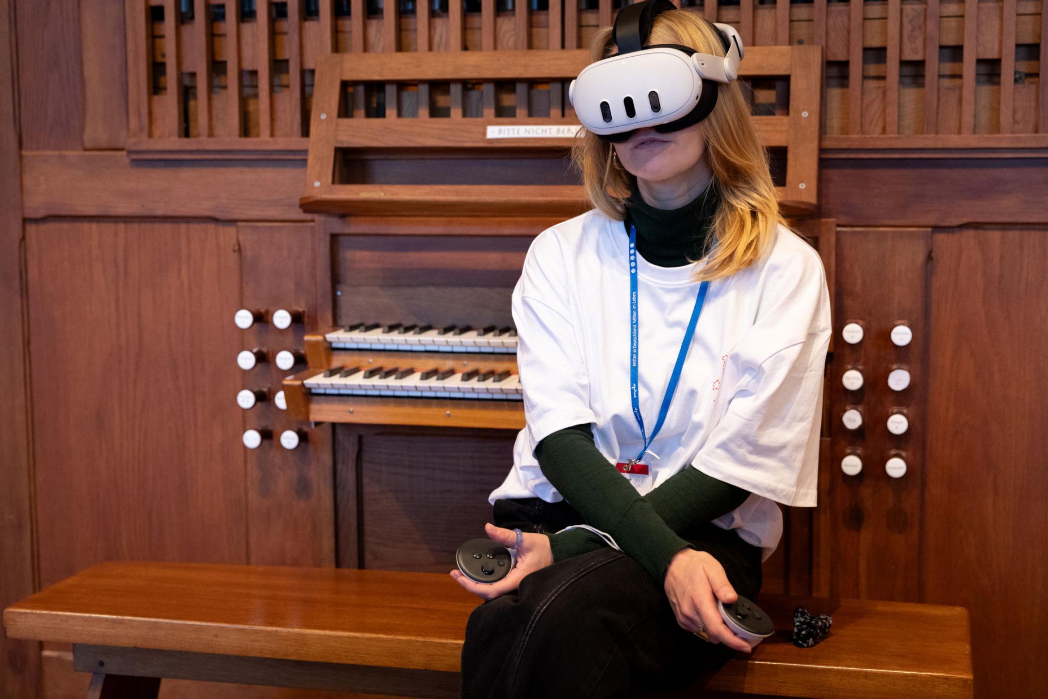 A woman sits in front of a huge church organ, wearing VR glasses, holding controllers in her hands and gazing into the distance at something only she can see. 