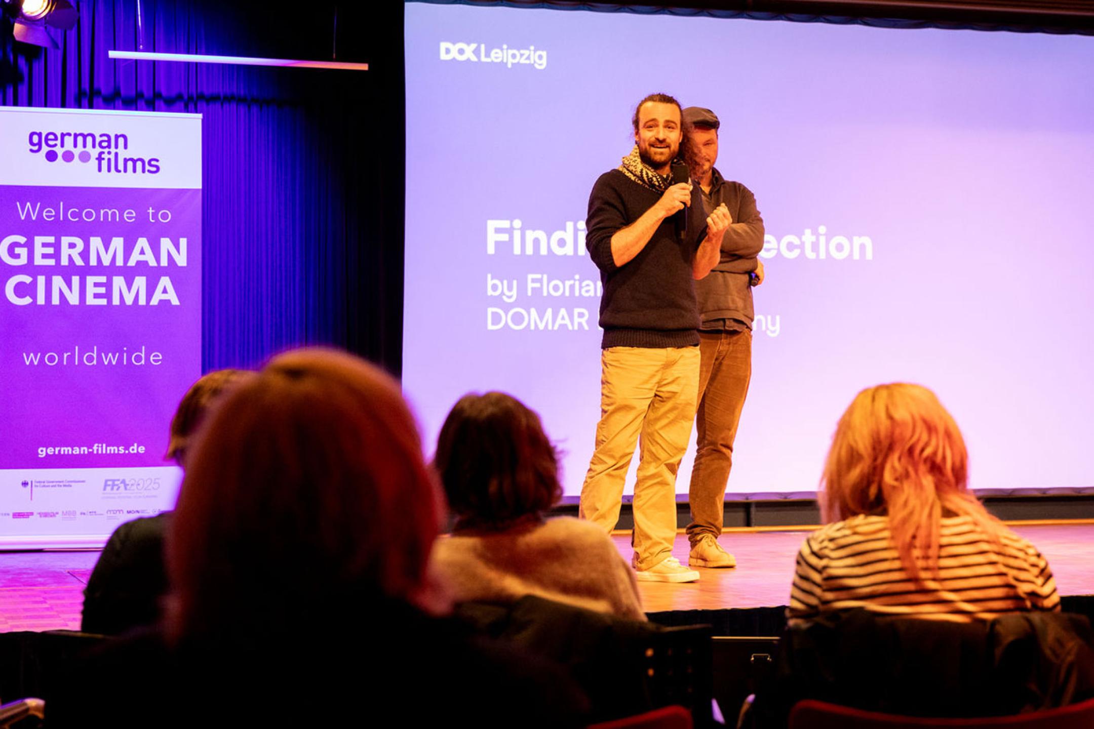 Two man with microphones on stage in front of an audience, the one closer to the camera is speaking and looking enthusiastically into the camera
