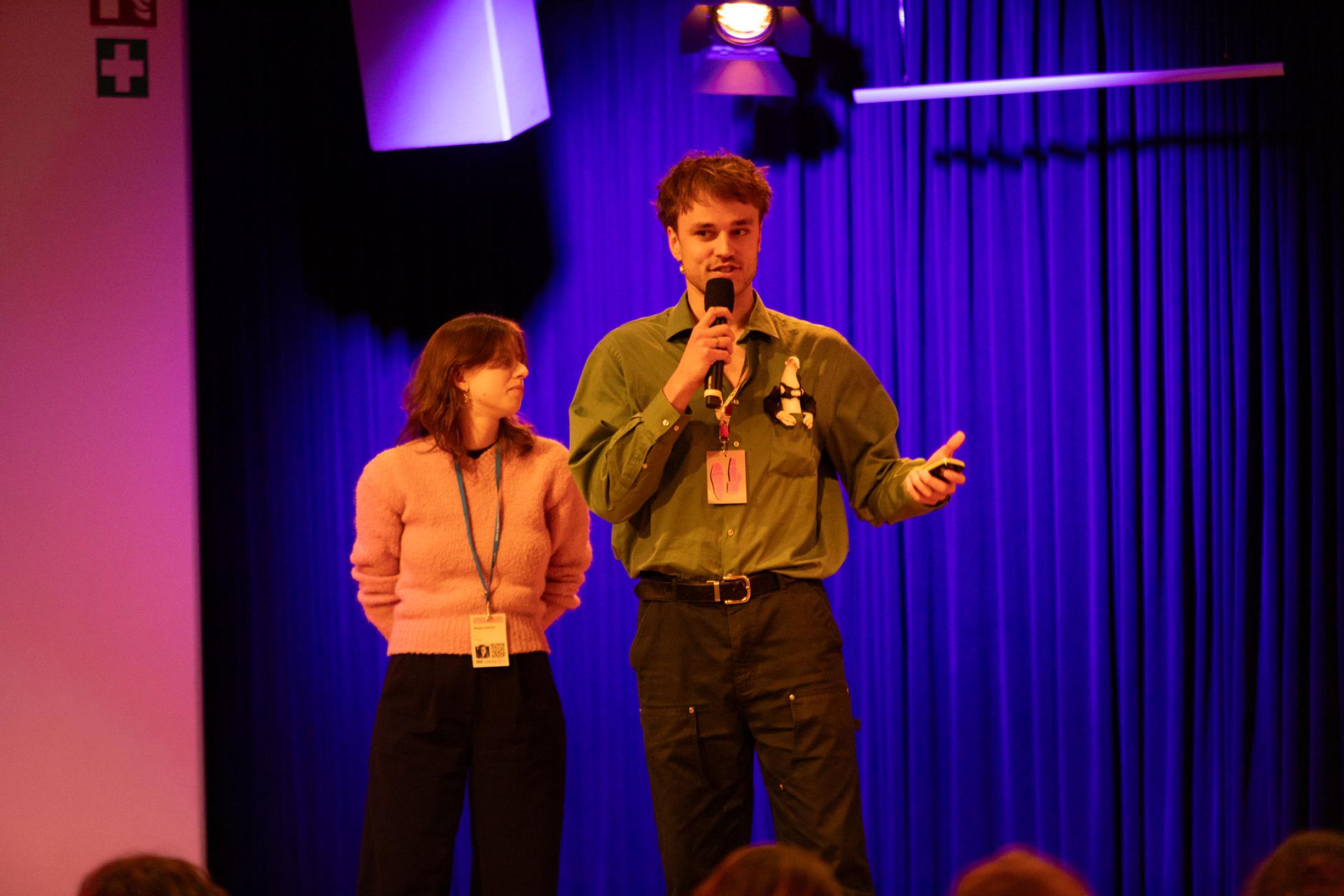 Zwei Personen auf der Bühne vor einem blauen Vorhang, eine von beiden spricht in ein Mikrophon 