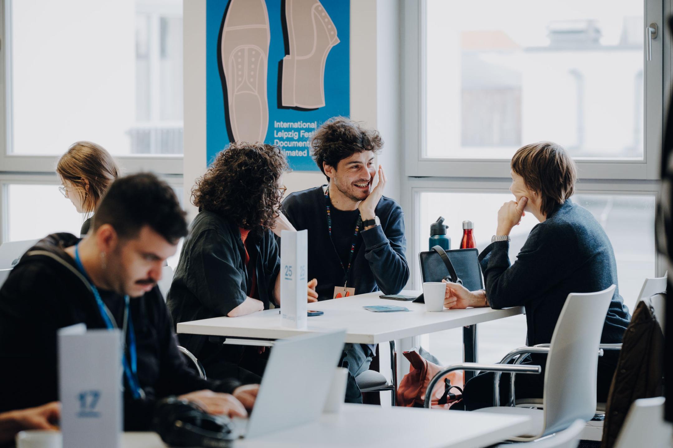 An einem Tisch vor einem DOK Industry Plakat sitzen drei Personen im Gespräch, einer von ihnen lacht gerade auf