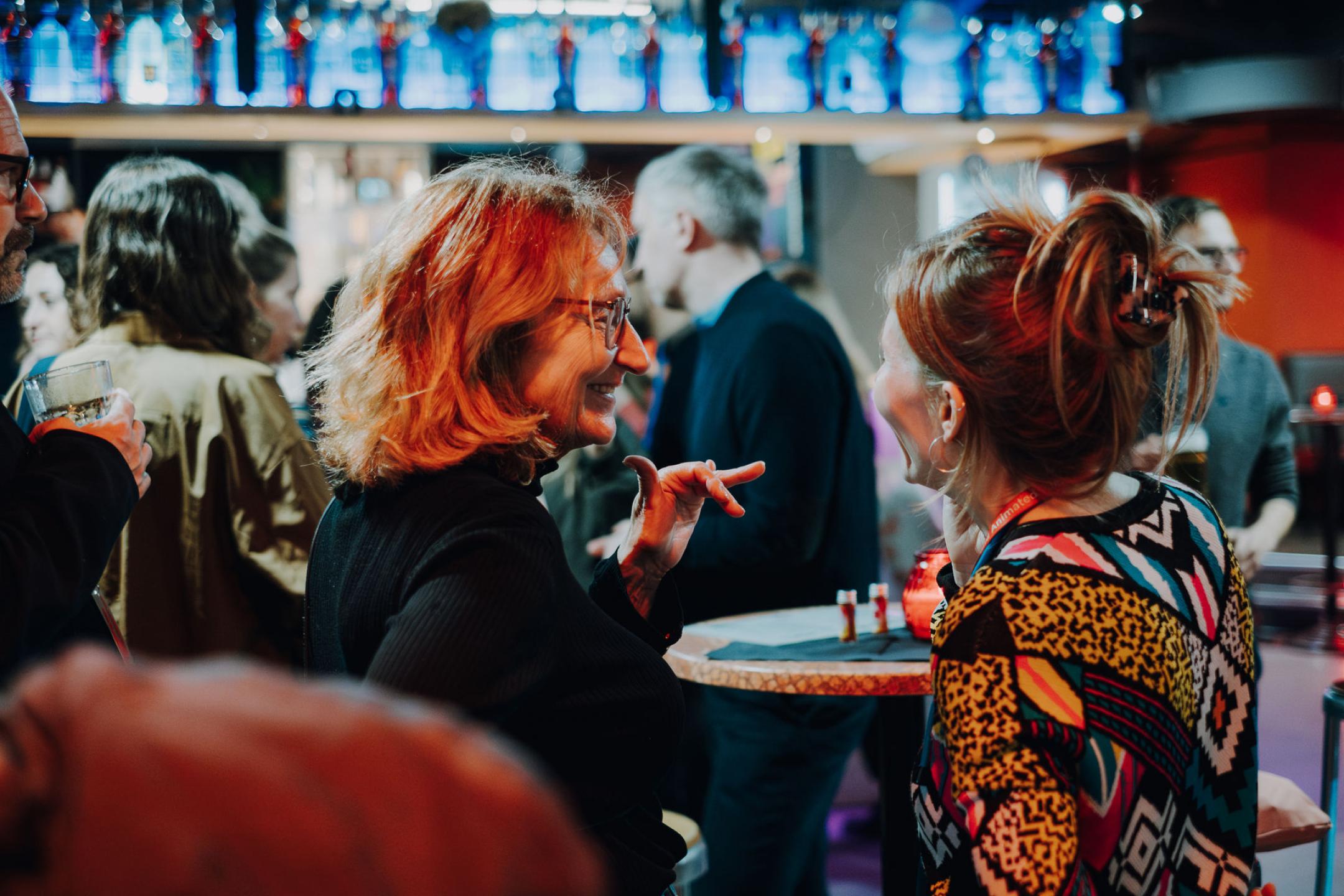Zwei Personen unterhalten sich angeregt in einer warm beleuchteten Bar voller Menschen