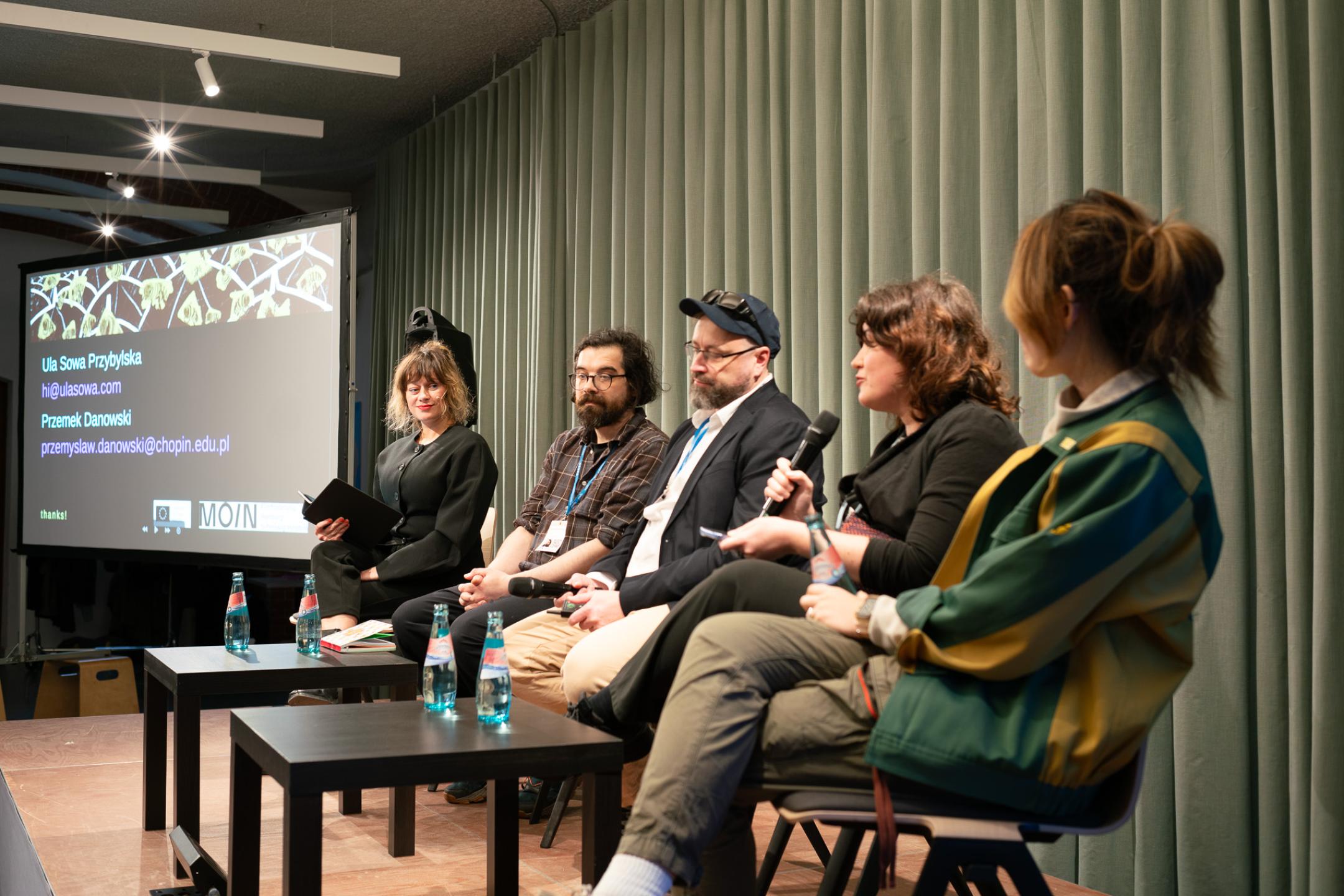 Fünf Menschen sitzen auf einer kleinen Bühne, eine Person spricht in ein Mikrophon