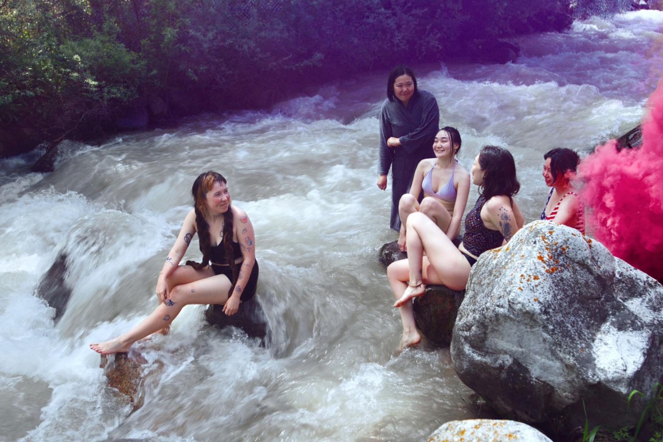 Ein paar junge Frauen sitzen lachend auf Steinen auf einem reißenden Fluss