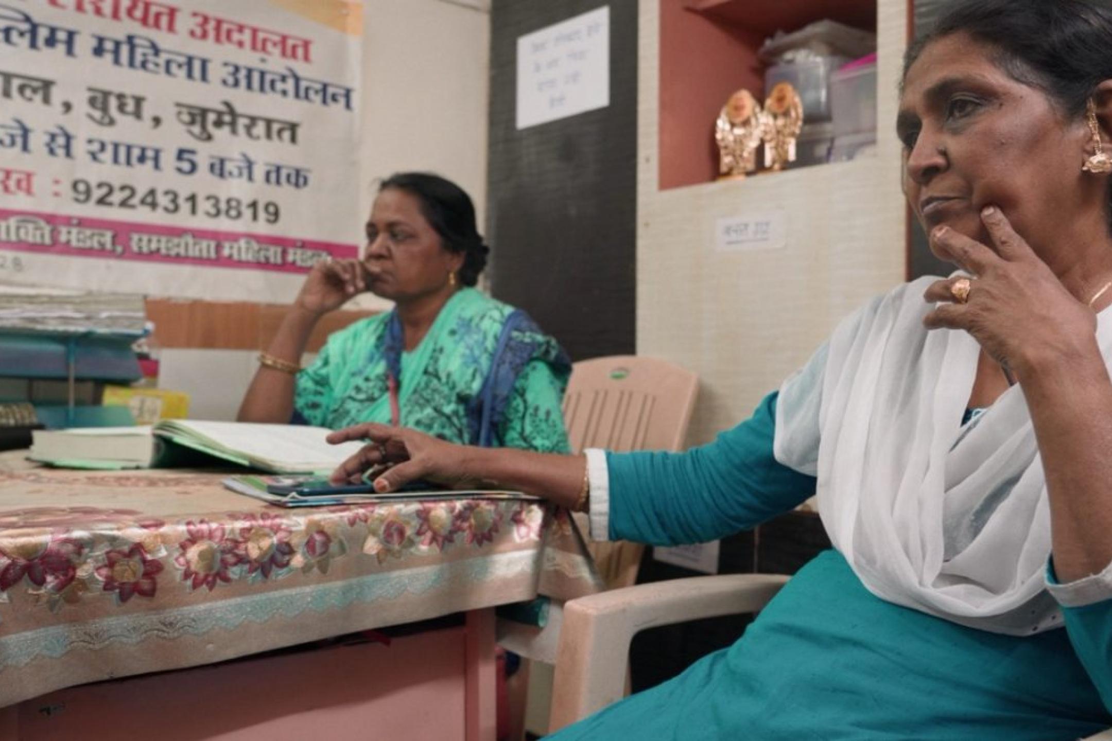 Zwei Frauen sitzen in einem kleinen Büro. Im Vordergrund sitzt eine Frau in türkis-weißer Kleidung nachdenklich auf einem Plastikstuhl und stützt ihr Kinn auf die Hand. Hinter ihr sitzt eine weitere Frau an einem Tisch mit Akten und telefoniert. An der Wand hängt ein Plakat mit Text in Hindi.