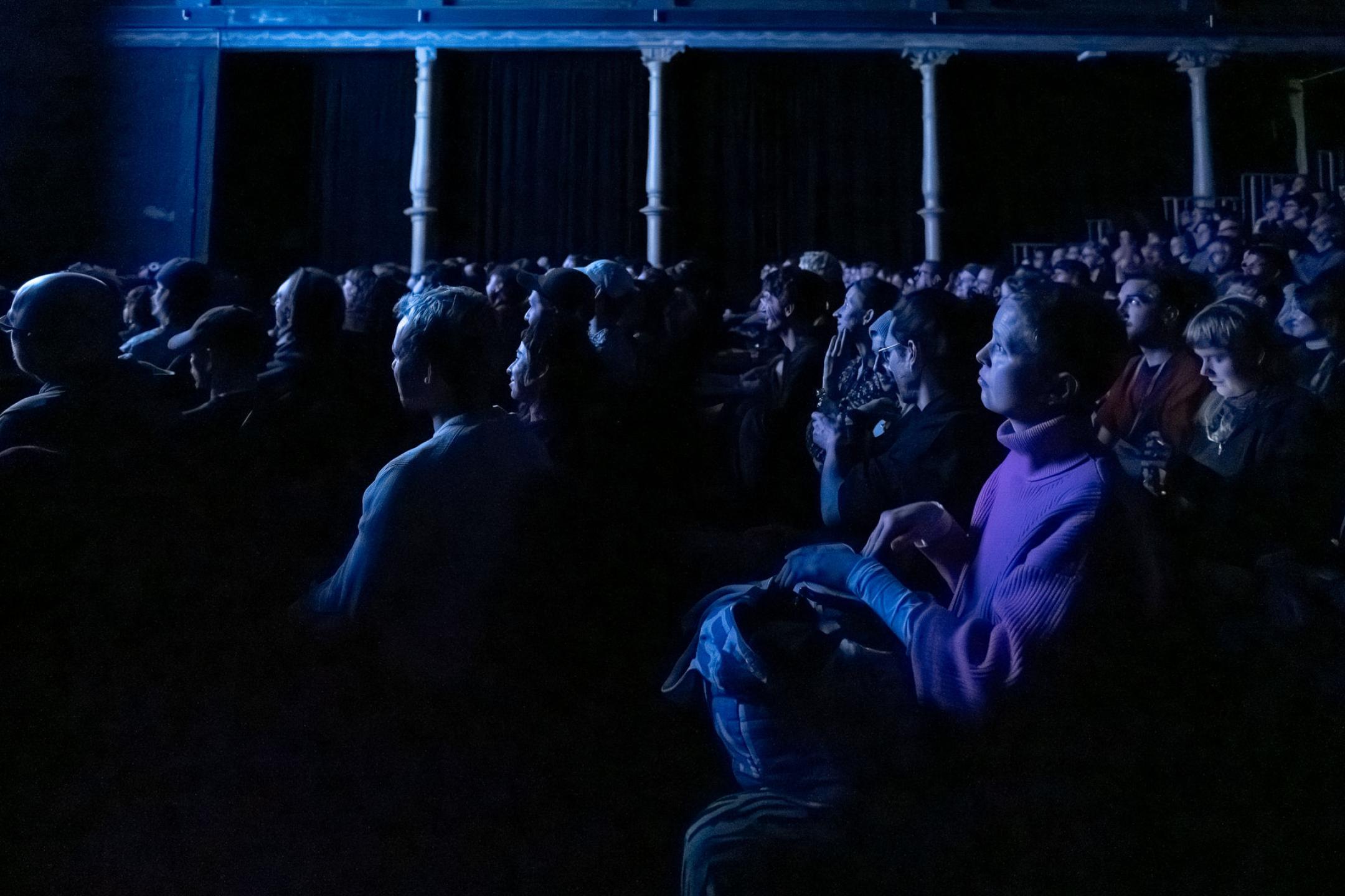 Zu sehen ist ein voller Kinosaal, mit Menschen, die richtung Leinwand schauen und in blaues Licht getaucht sind.