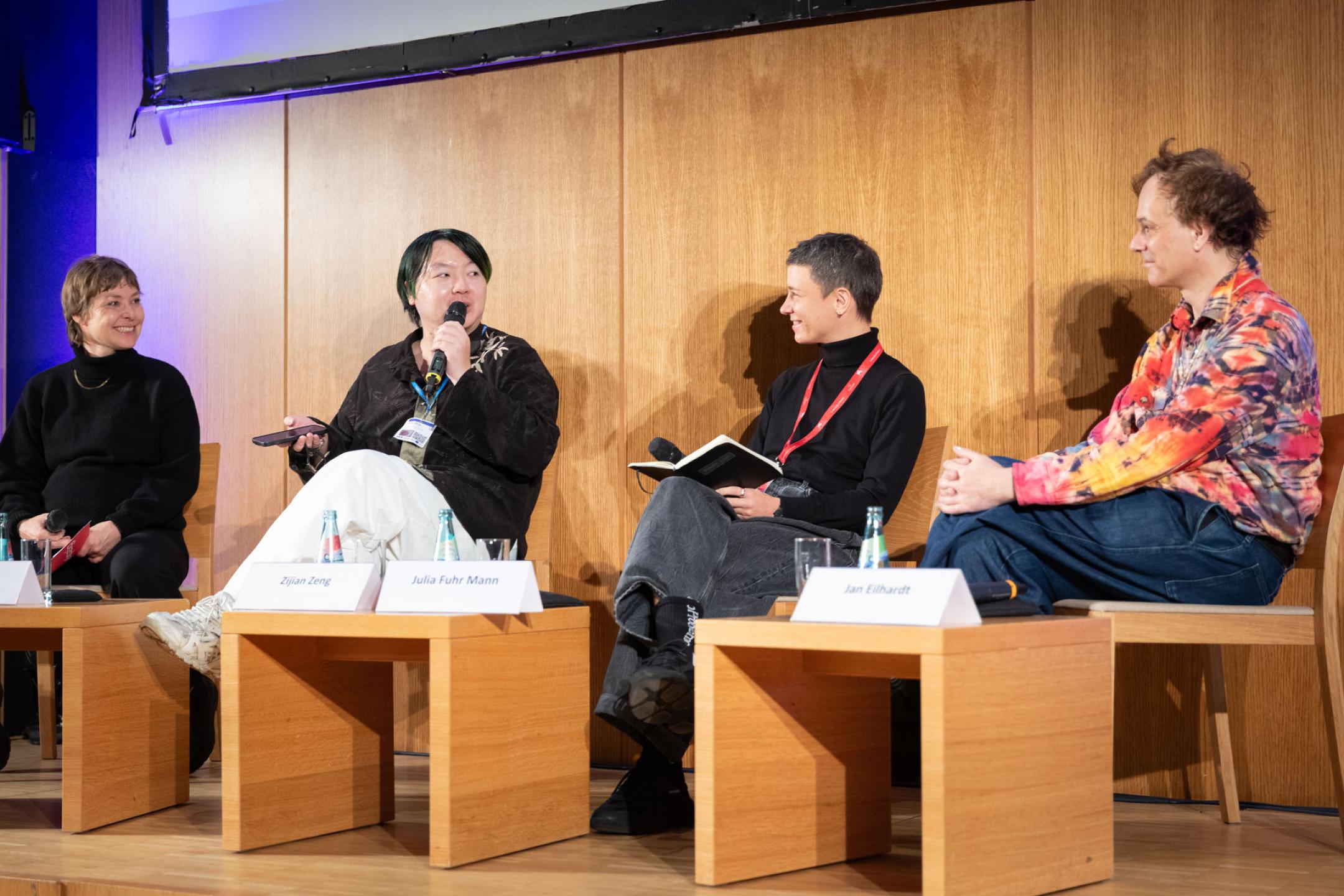 Vier Personen sitzen auf einer Bühne zusammen und führen eine Panel Diskussion. Hinterihnen ist eine hölzerne Wand und vor ihnen stehen kleine Holztische mit Getränken und Namensschildern.