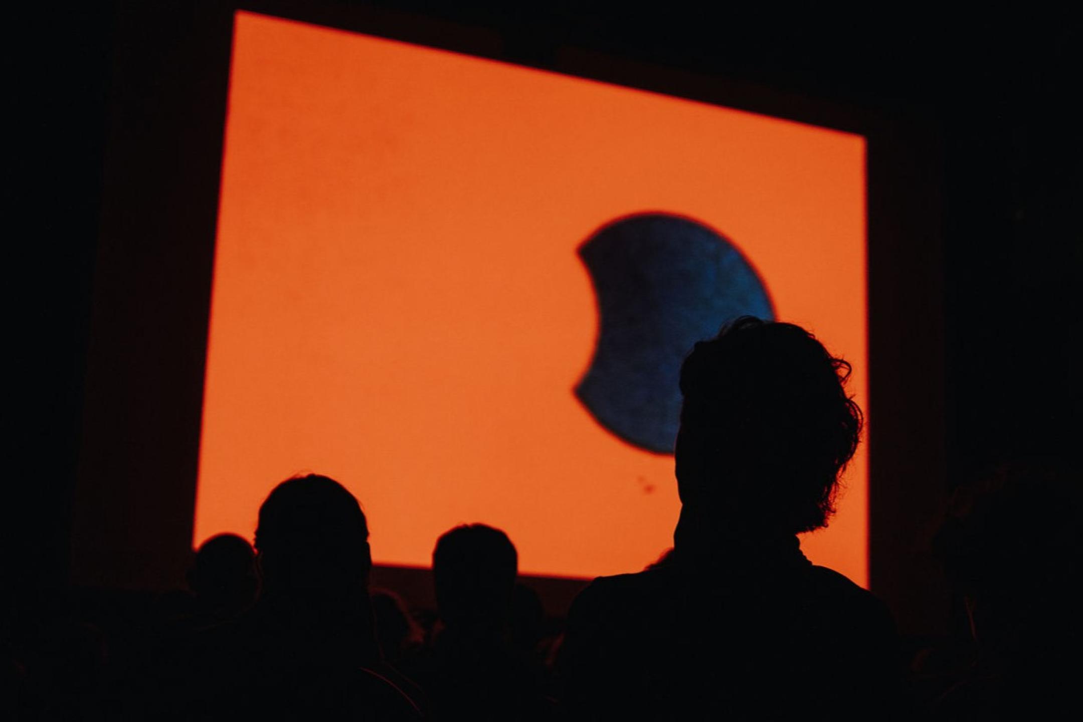 Die Kinoleinwand zeigt einen orangenen Hintergrund auf dem ein schwarzer Kreis zusehen ist, der jedoch auf der linken Seite nicht ganz voll ist und an Pac-Man erinnert. Davor sind nur die dunklen Silhouetten der Zuschauenden zu erkennen. 