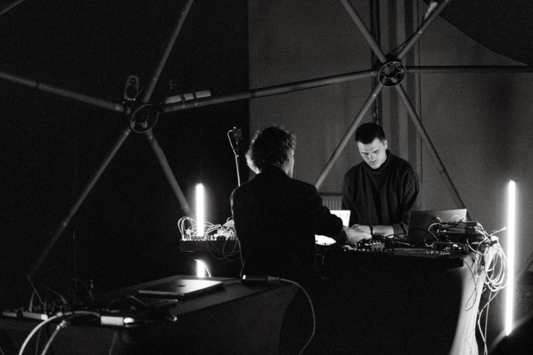 Schwarz-weiß-Foto von zwei Menschen an einem DJ Pult, um sie herum ist eine Rohrkonstruktion aufgebaut und längliche LED-Leuten