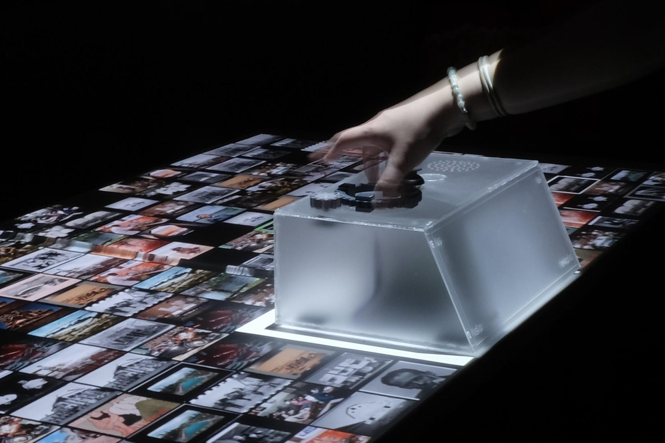 Eine Hand greift nach einem halbtransparenten Plastikbox, die auf einer Fläche voller Fotos steht