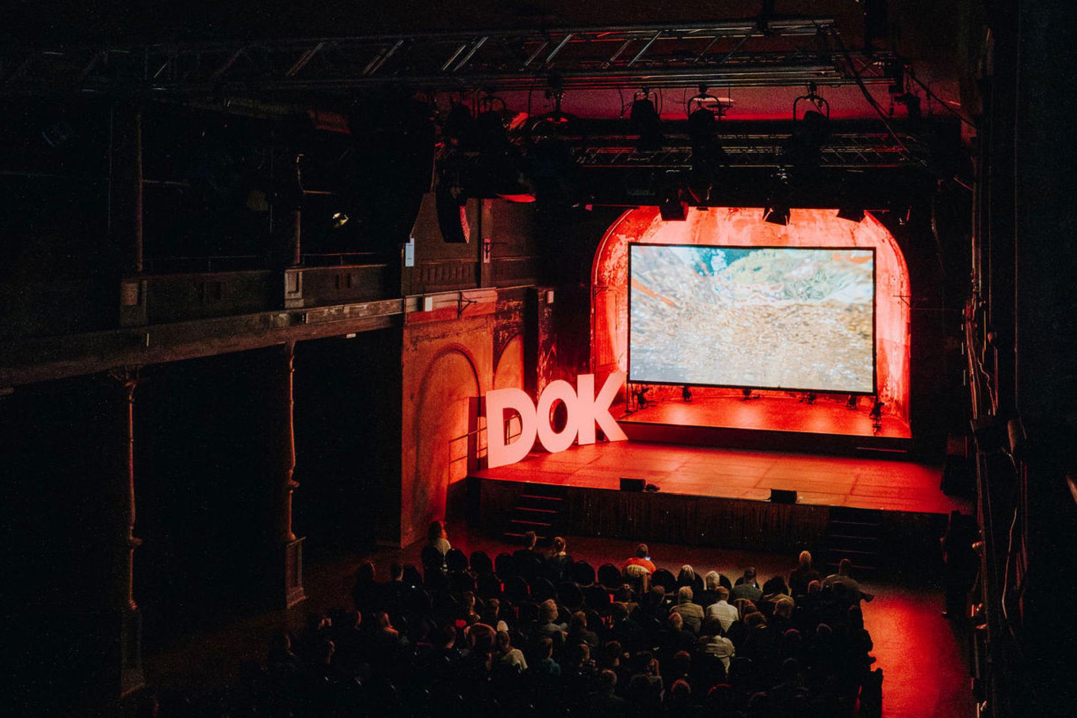 Ein Kino mit einer hellen Leinwand und riesigen Papierbuchstaben, die auf der Bühne das Wort „DOK“ bilden