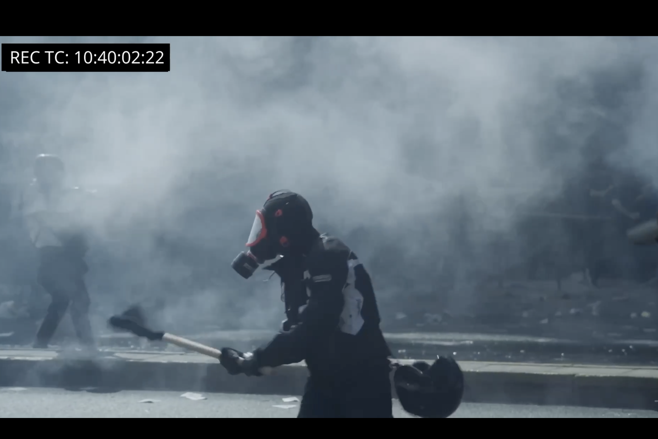 Eine Person die eine Gasmaske trägt, läuft durchs Bild. Im Hintergrund ist Rauch zu sehen und es liegen Gegenstände herum. Es scheint eine Momentaufnahme von einer Demo oder Ausschreitung zu sein.