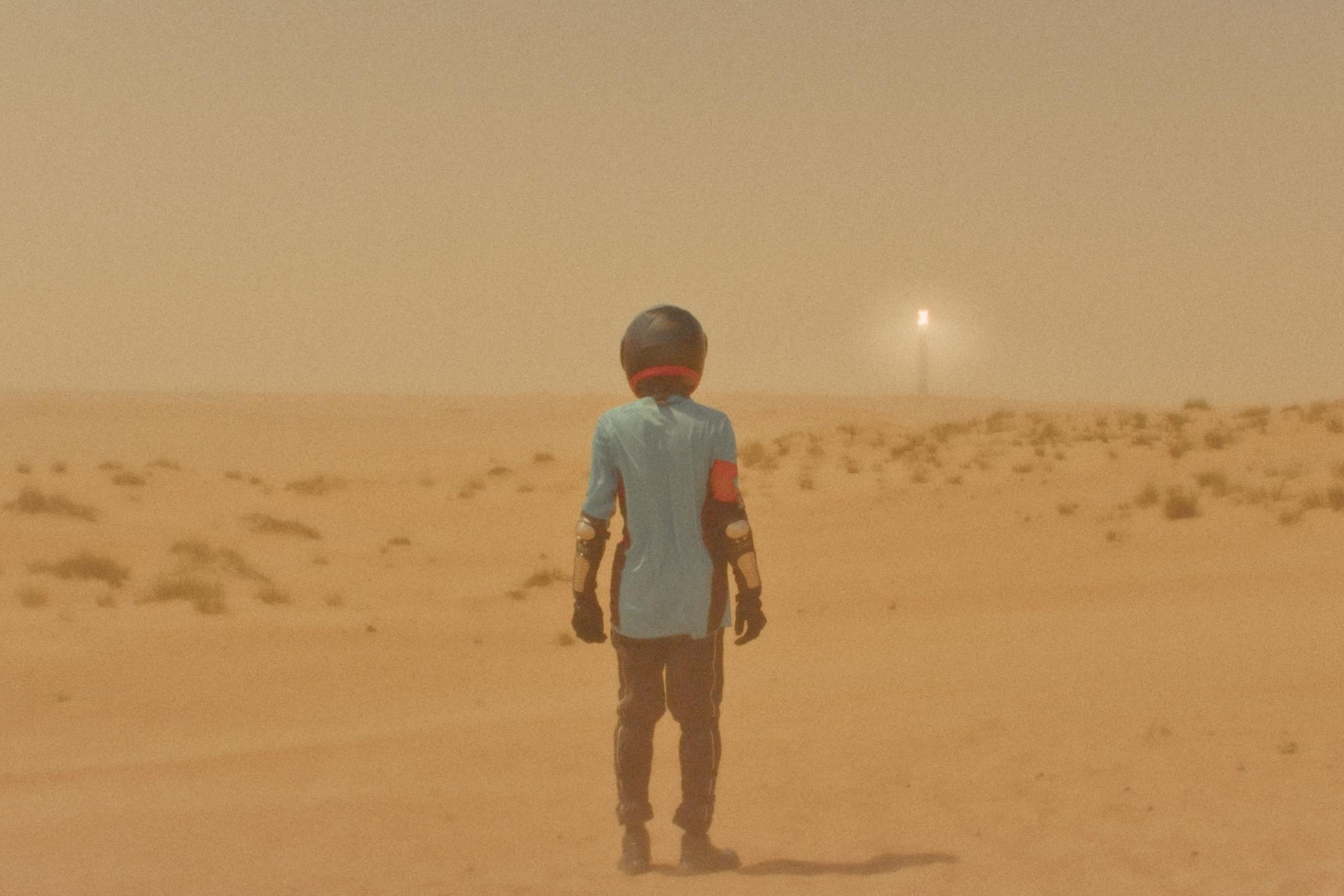Gelber Nebel, in einer Wüstenlandschaft steht eine Person mit Motorradhelm, sehr dunstige Atmosphäre und in der Ferne ein Laternenlicht
