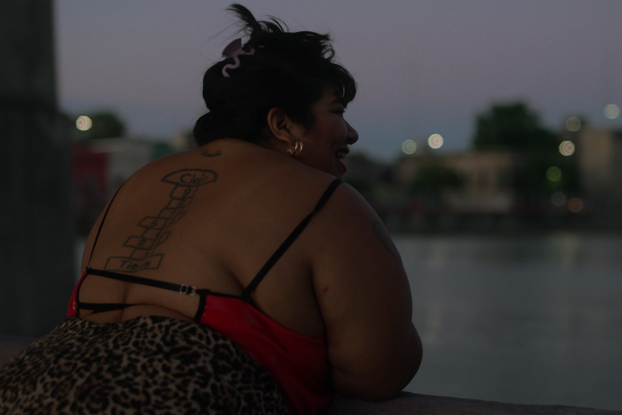 Eine dicke Frau lehnt in der Abenddämmerung über ein Geländer. Sie trägt eine Leoprint Hose und ein Oberteil mit tiefem Rückenausschnitt, sodass ihr Tattoo auf der Wirbelsäule sichtbar ist.