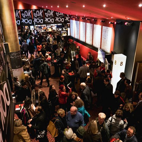 Foyer des Cinestar Leipzig mit vielen Besucherinnen und Besuchern, die ins Kino strömen, viele DOK Leipzig Plakate im Hintergrund.