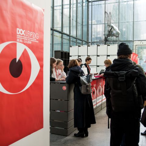 Leute in der Schlange im Gästebüro warten darauf akkreditiert zu werden. Links an einer Wand hängt ein großes rotes Plakat mit dem Festivalmotiv.