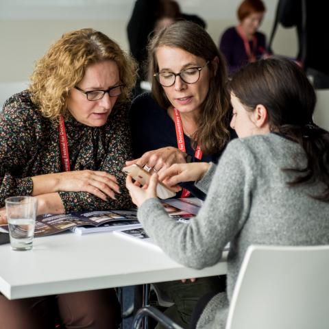 Drei weibliche Personen sitzen an einem Tisch und schauen gemeinsam auf ein Smartphone-Display.
