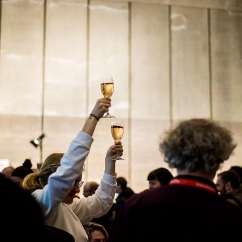 A woman with two glasses of wine in the middle of a crowd.