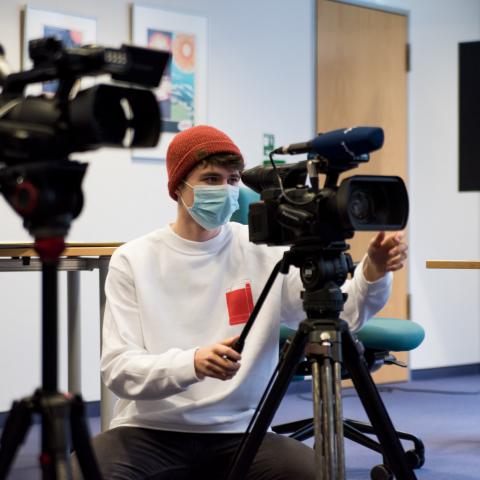 Ein Jugendlicher mit Mundschutz blickt durch den Sucher einer Videokamera.