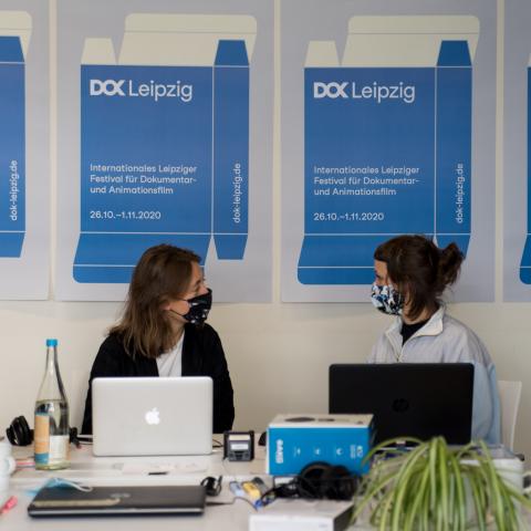 Eine Reihe DOK Industry Plakate an der Wand, davor sitzen zwei Frauen mit Maske vor Laptops, die einander anschauen