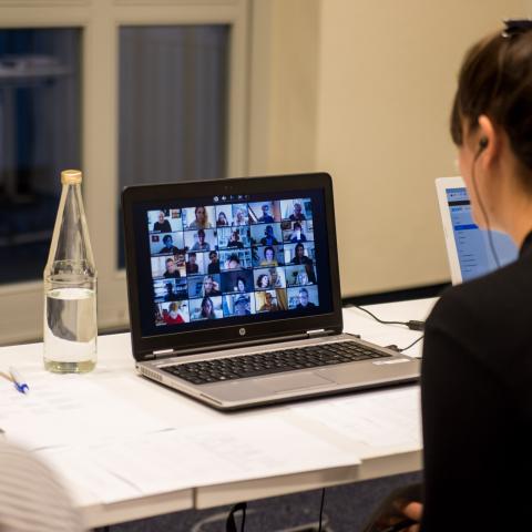 A woman is sitting in front of her laptop in which a zoom conference with many participants can be seen