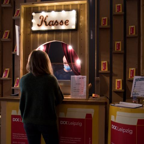 Eine Frau steht an einer Kinokasse, die großflächig von DOK Leipzig Plakaten umrahmt ist.