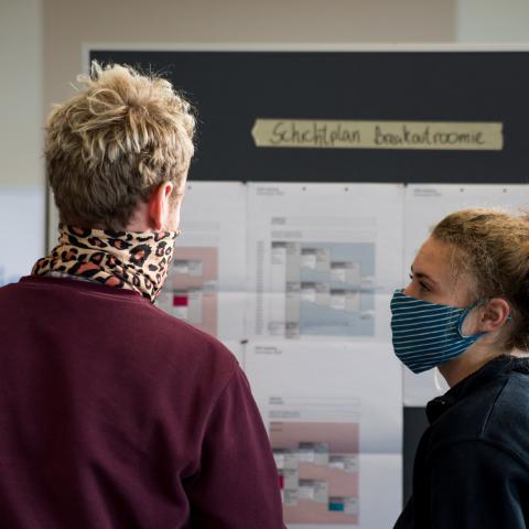 Eine DOK-Mitarbeiterin und ein DOK Mitarbeiter – beide mit Mundschutz – unterhalten sich vor einer Wandtafel mit einem Schichtplan. Über dem Plan die Überschrift "Breakoutroom"