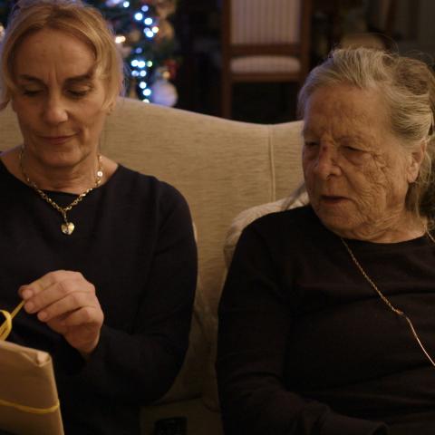Szene aus dem Film "The Blunder of Love": Eine jüngere und eine ältere Frau sitzen nebeneinander auf einem Sofa. Die jüngere Frau (links) packt ein Geschenk aus, die ältere schaut dabei zu.