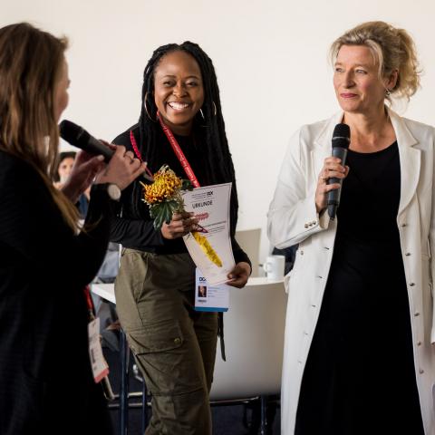 One smiling black woman with an award and 2 white women with microphone looking at her.
