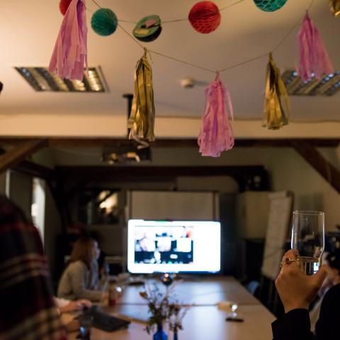 Zwei Personen, die ein Glas in der Hand halten, blicken auf einen hell erleuchteten Monitor. Über ihnen hängt eine Partygirlande, das Licht ist schummrig.
